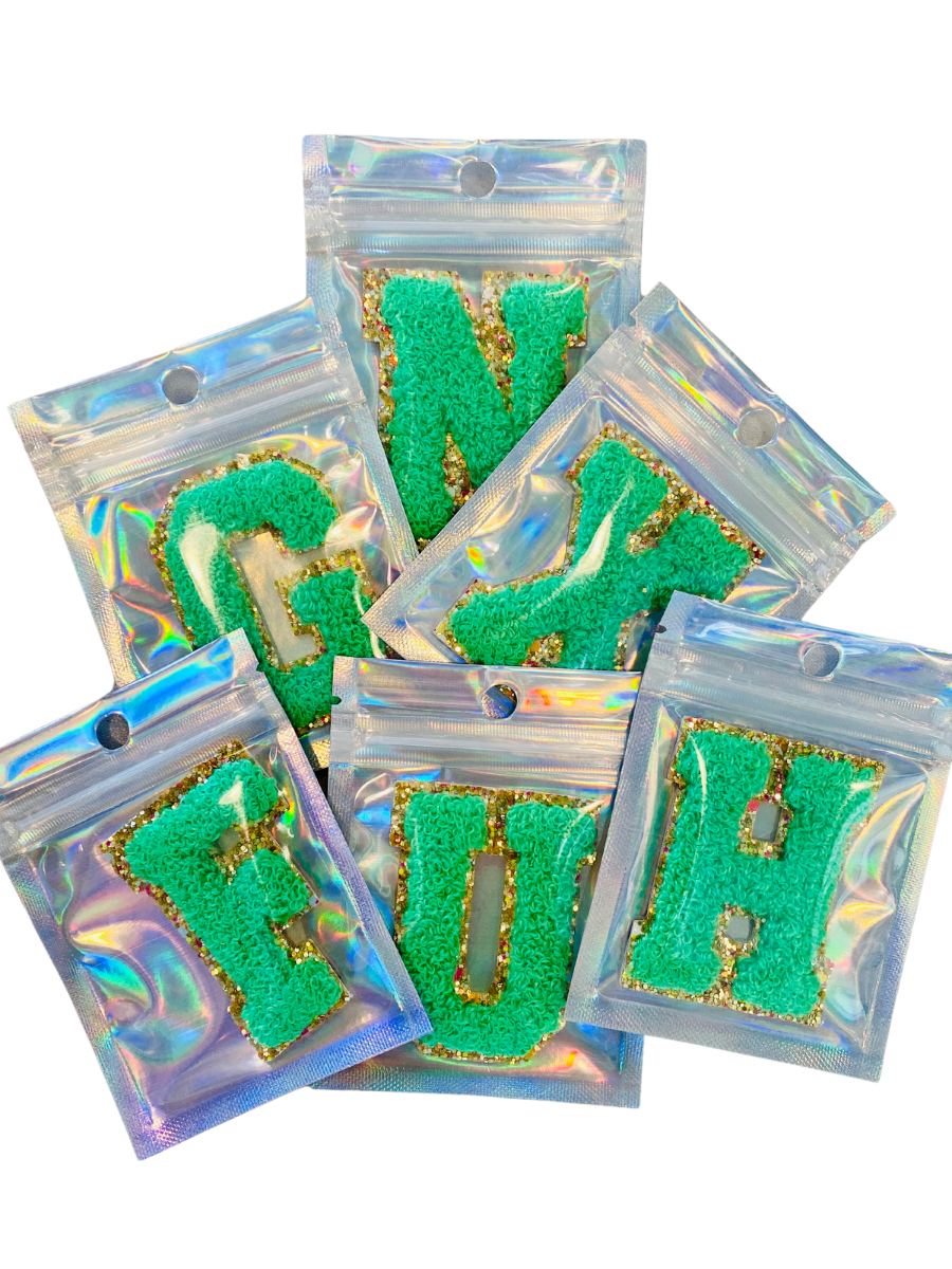 Six green chenille letter patches with gold glitter borders are individually packaged.