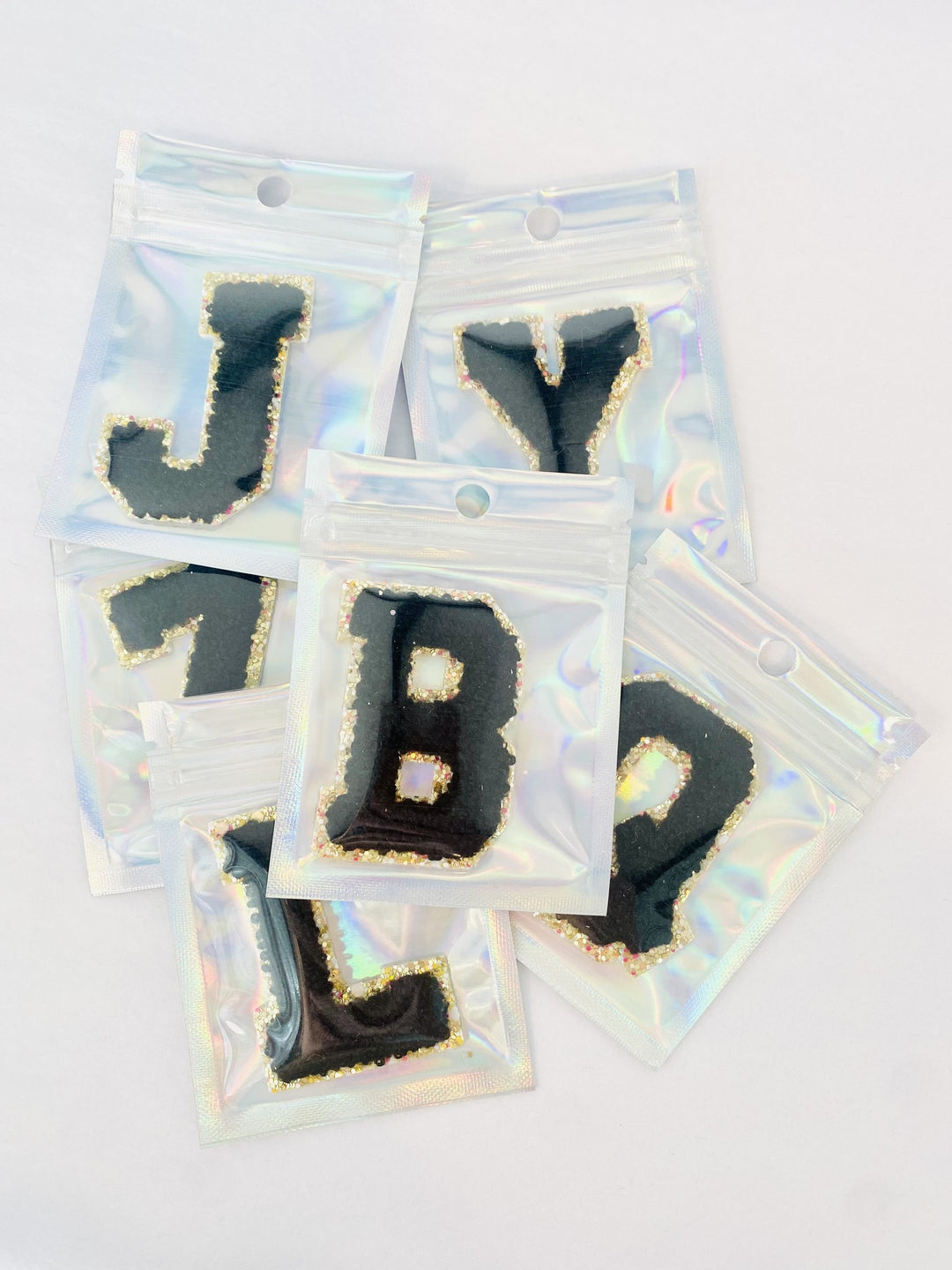 A pile of black chenille letter patches with glitter borders in clear holographic packaging.