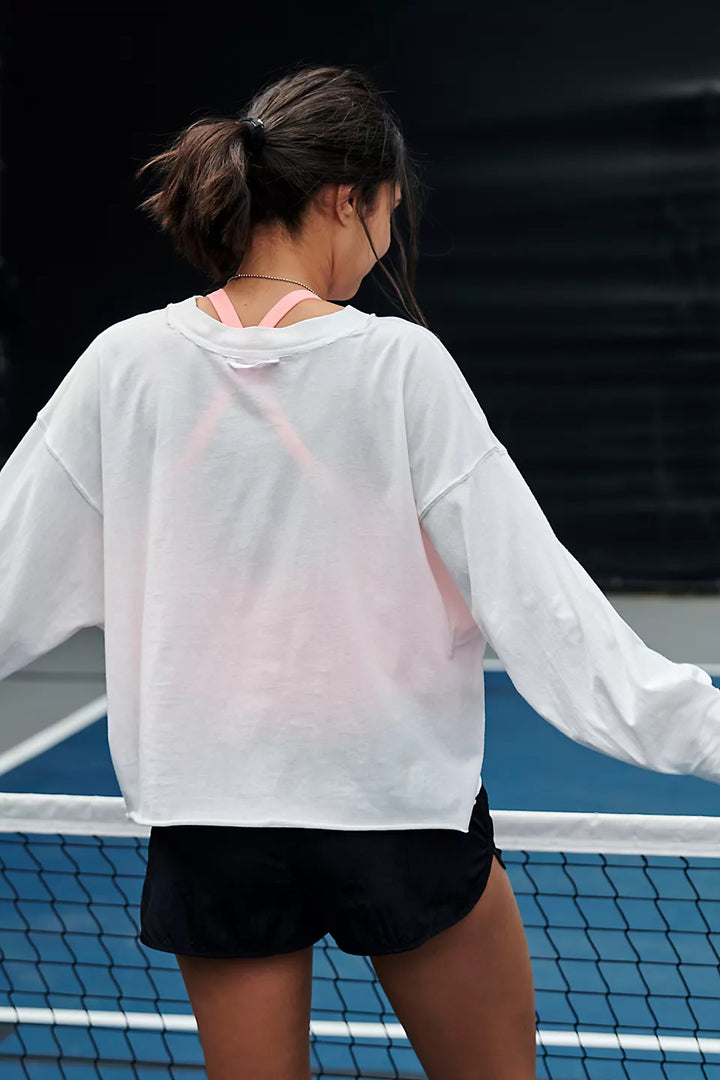 A woman from behind wears a white long-sleeve top, pink sports bra, and black shorts on a tennis court.