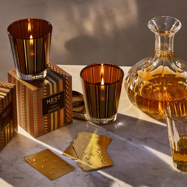 Two lit Nest New York Hearth candles in striped glass vessels sit next to a decanter and playing cards.