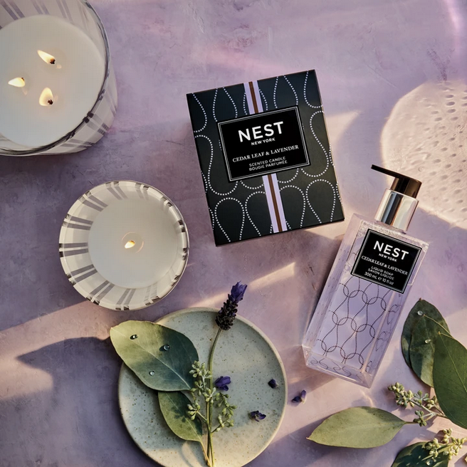 A Nest New York Cedar Leaf & Lavender 3-wick candle, a single-wick candle, and liquid soap are arranged with lavender and eucalyptus leaves.