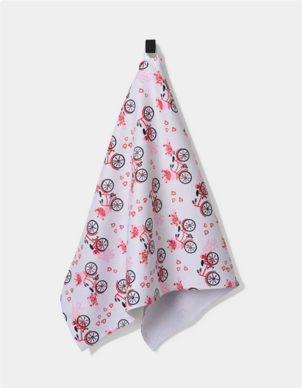 White tea towel with a pattern of red bicycles and pink hearts, hanging from a black hook.