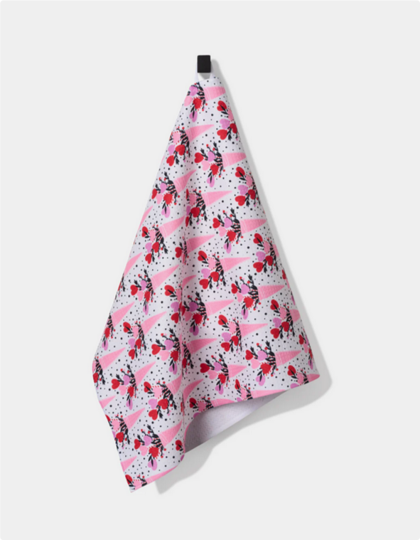 A white tea towel with a repeating pattern of pink cones filled with red and pink hearts hangs from a black hook.