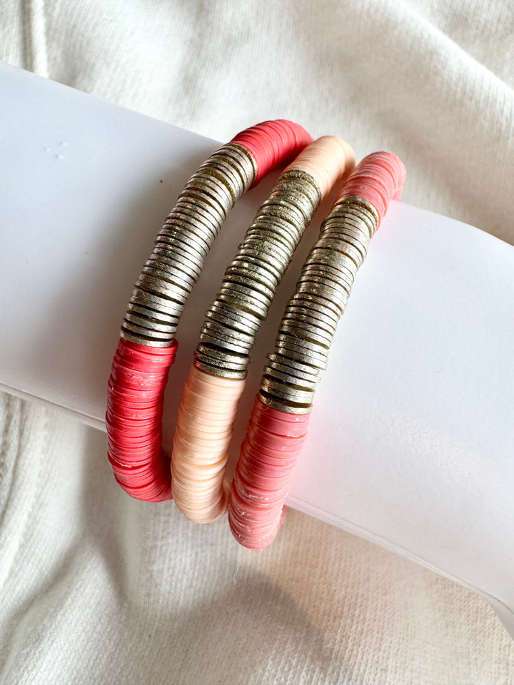 Three flat bead bracelets in shades of pink and gold are stacked on a white surface.