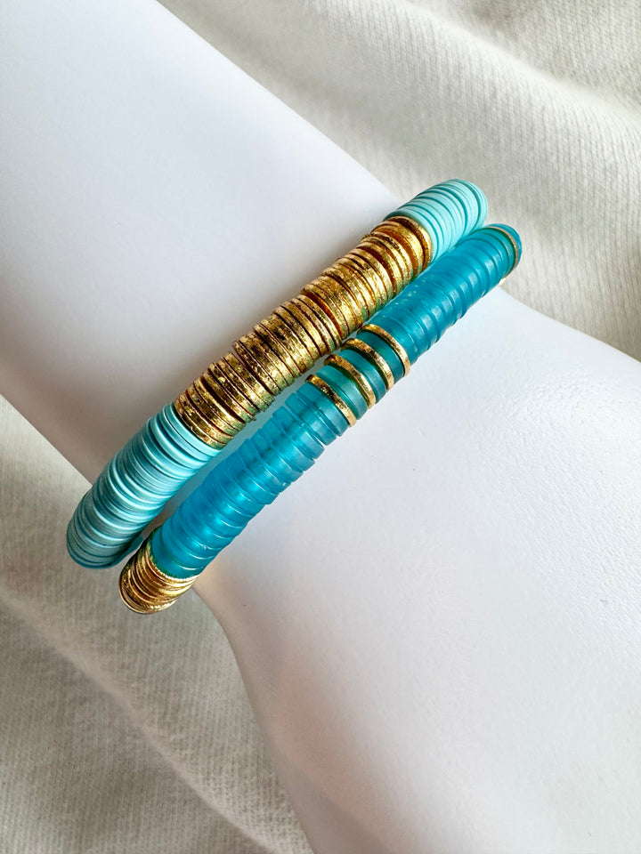 Two flat bead bracelets, one gold and one blue, are stacked on a white surface.