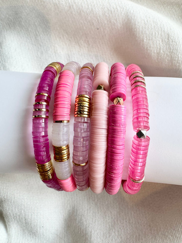 Seven flat bead bracelets in shades of pink, purple, and white with gold accents.