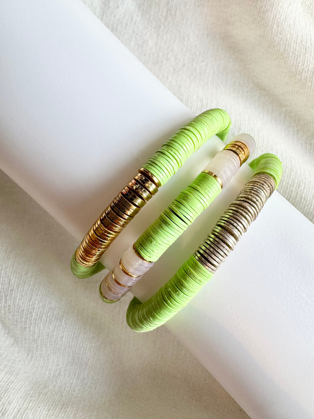 Three flat bead bracelets, one gold, one silver, and one mixed, rest on a white surface.