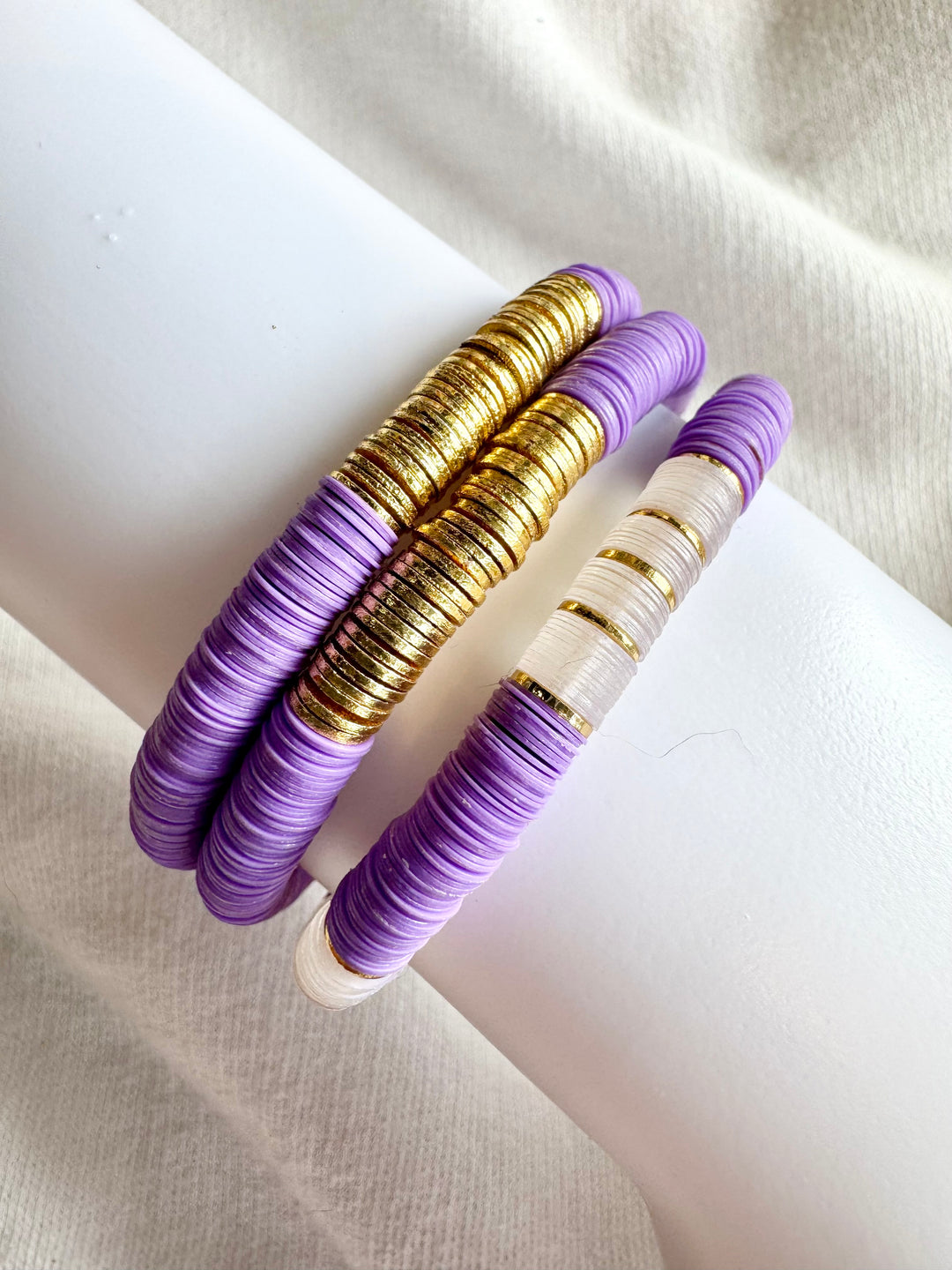 Three flat bead bracelets, two purple and gold, one purple, white, and gold.