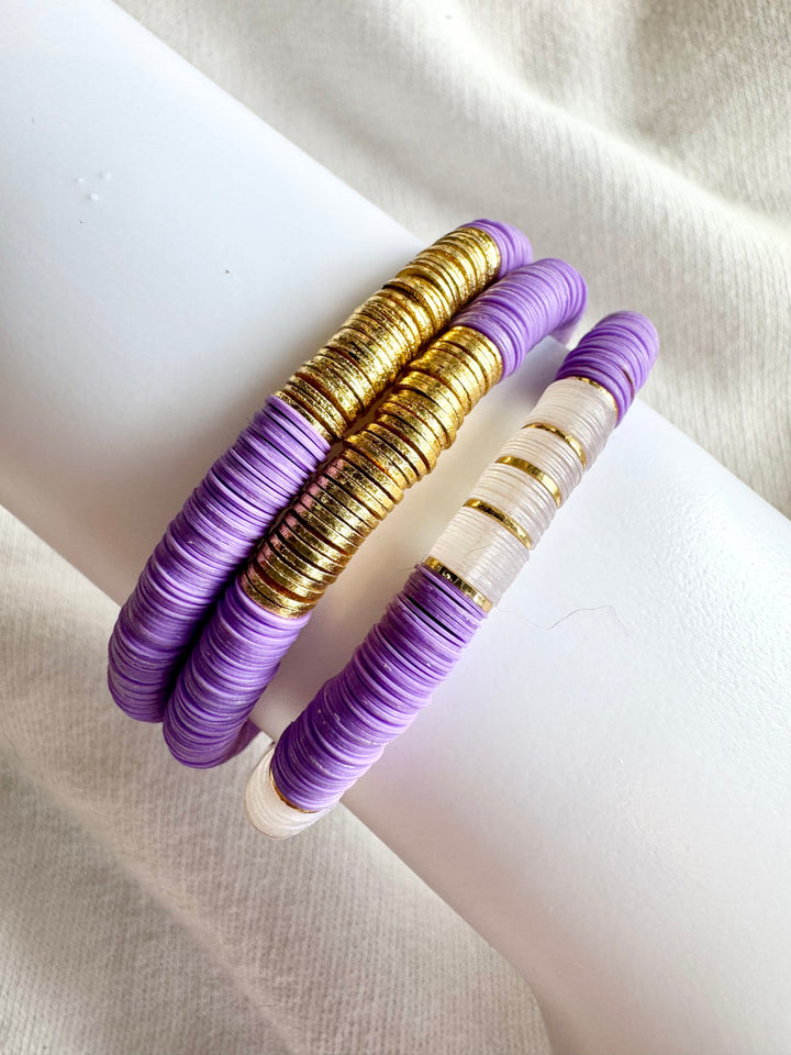 Three flat bead bracelets, two purple and gold, one purple, white, and gold.