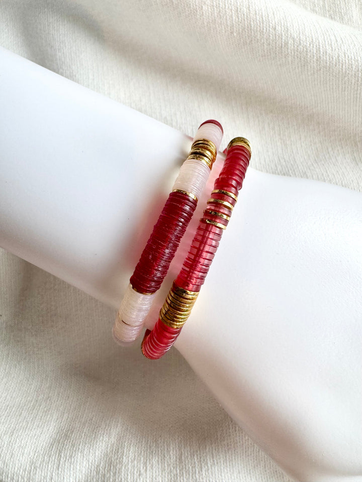 Two red and white flat bead bracelets with gold accents are displayed on a white surface.