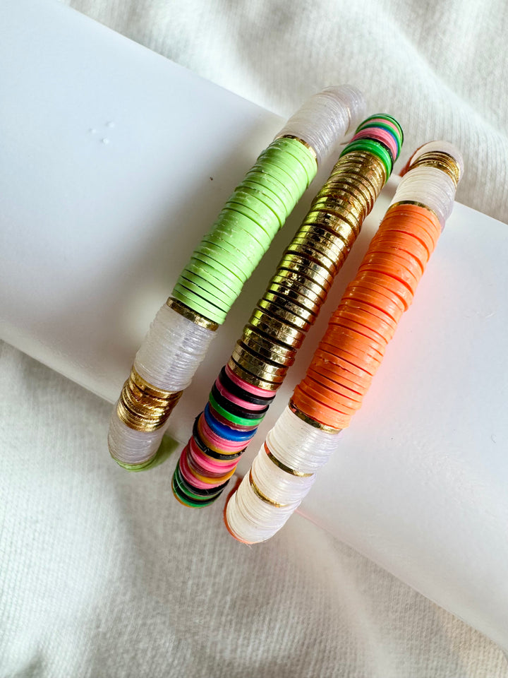Three flat bead bracelets, one green, one gold, and one orange, rest on a white surface.