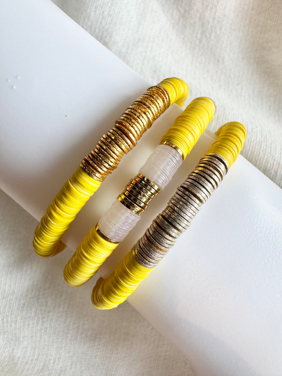 Three flat bead bracelets, one gold, one white and gold, and one silver, on a white surface.
