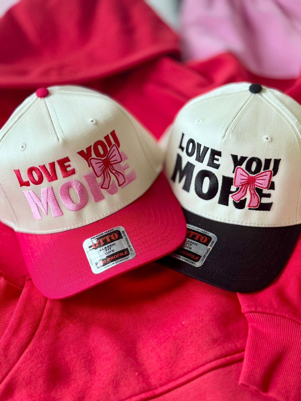 Two embroidered hats that say "Love You More" with pink bows.