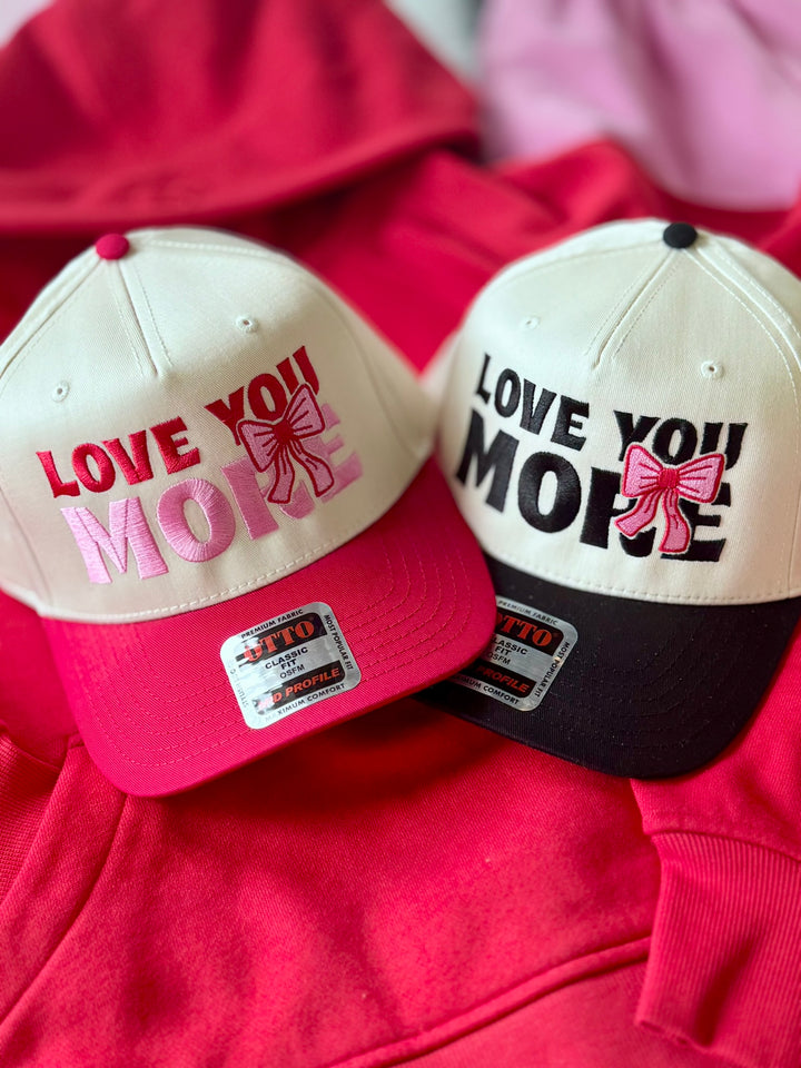 Two embroidered hats that say "Love You More" with pink bows.