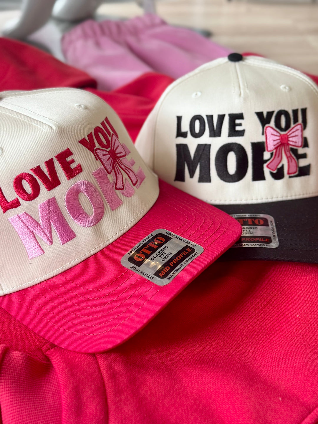 Two embroidered hats, one with "LOVE YOU MORE" in pink and red, the other with "LOVE YOU MORE" in black and pink.