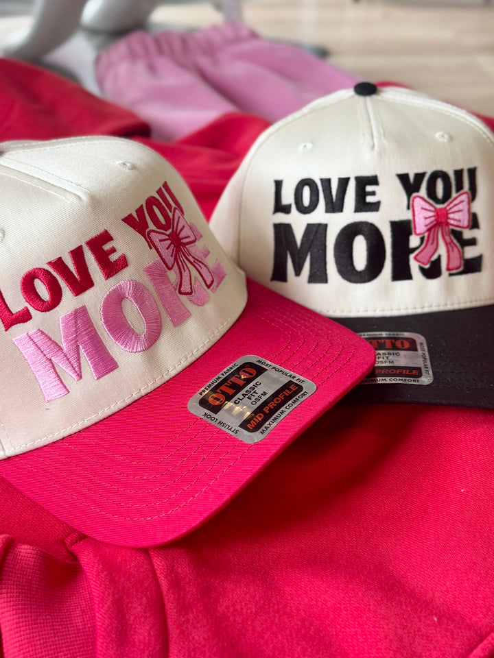 Two embroidered hats, one with "LOVE YOU MORE" in pink and red, the other with "LOVE YOU MORE" in black and pink.