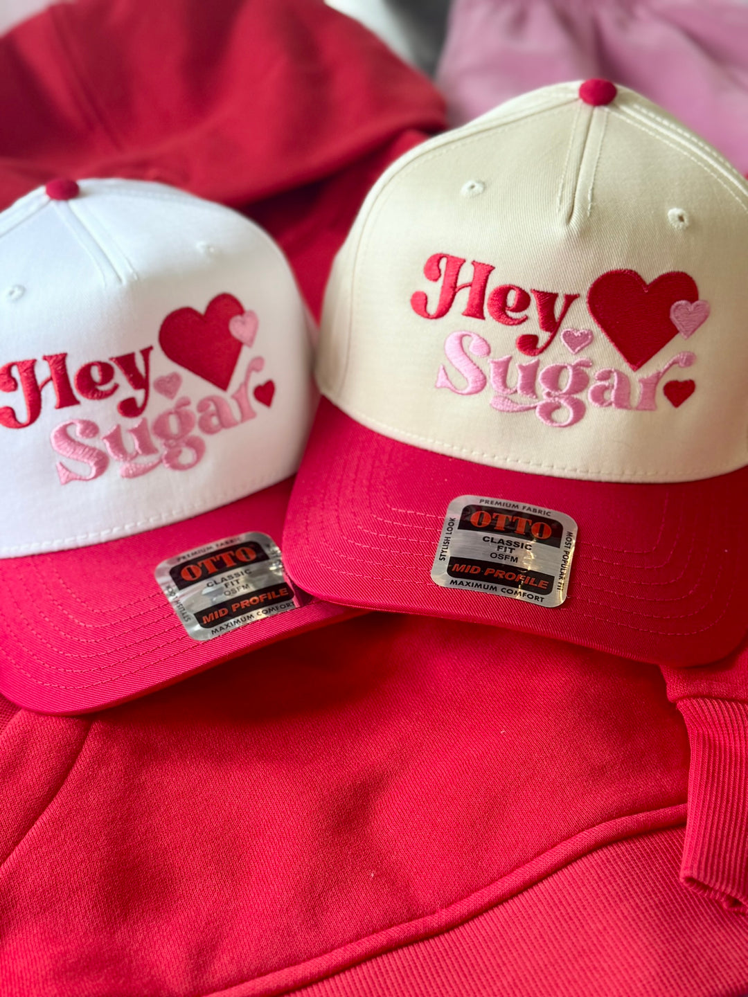 Two embroidered hats with "Hey Sugar" and hearts on a red background.