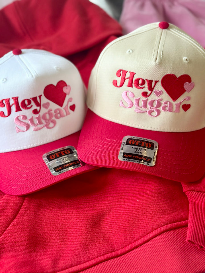 Two embroidered hats with "Hey Sugar" and hearts on a red background.