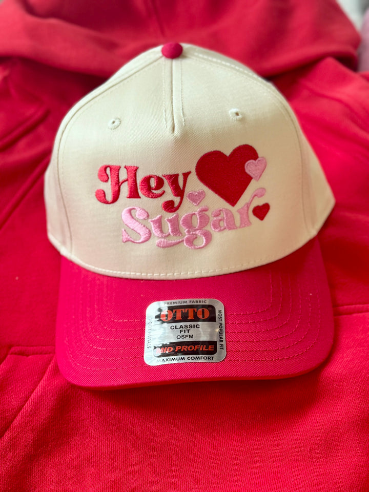 A cream and pink baseball cap with "Hey Sugar" embroidered in pink and red.