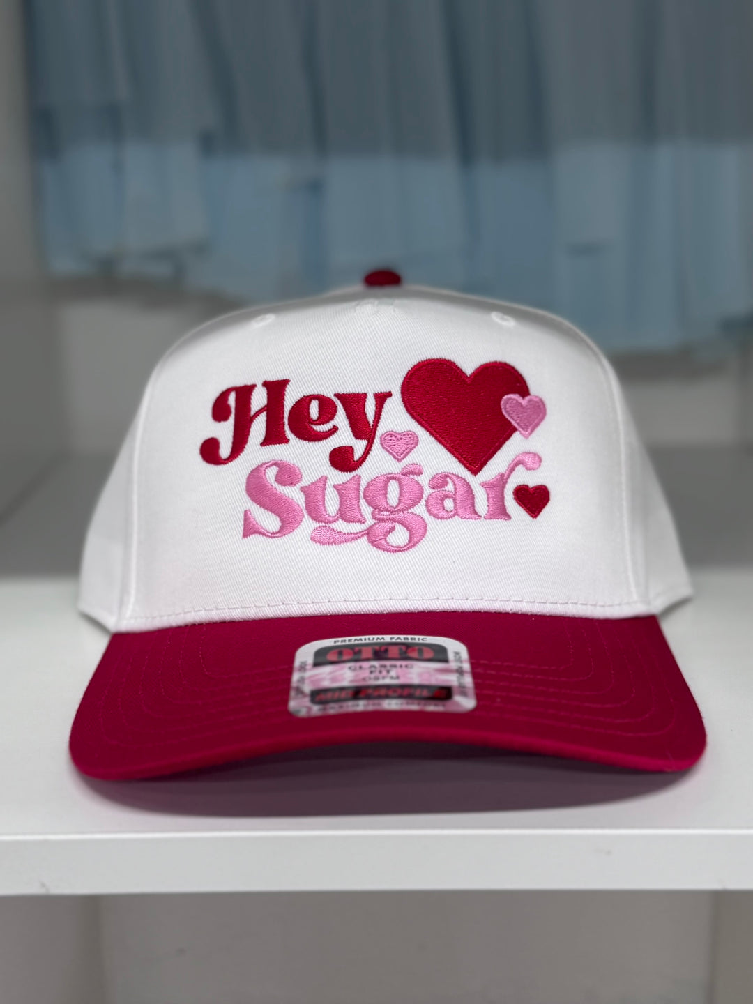 A white trucker hat with a pink brim and "Hey Sugar" embroidered in pink and red with hearts.