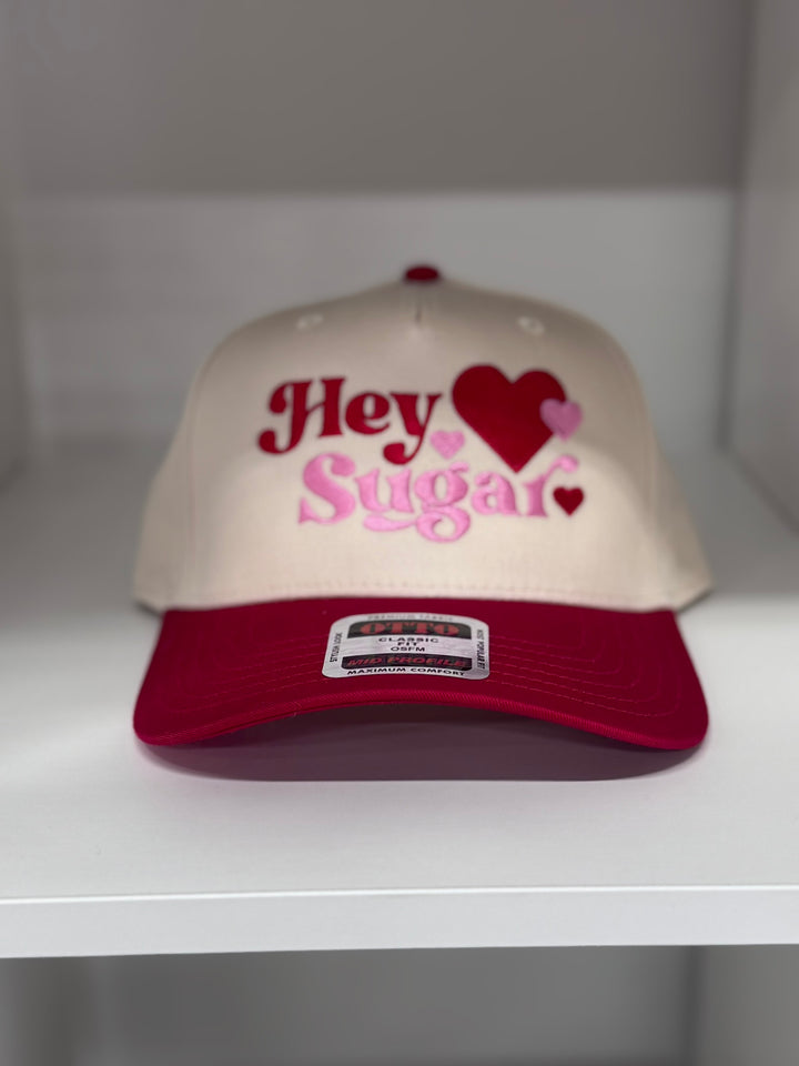 A cream baseball cap with red and pink "Hey Sugar" embroidery and hearts.