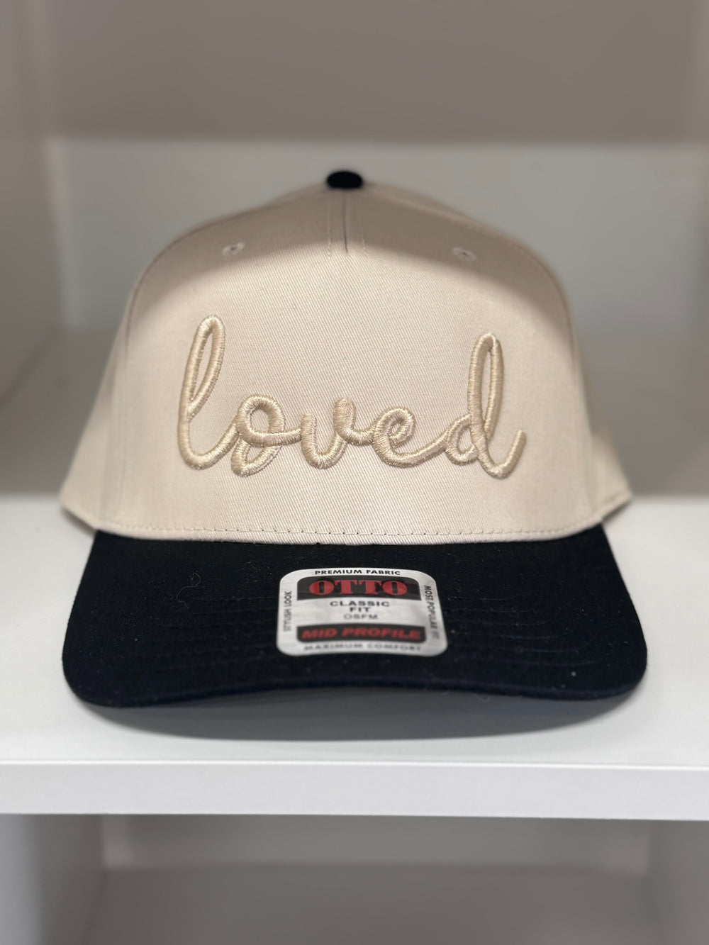 A tan baseball cap with "loved" embroidered in cursive on the front and a black bill.