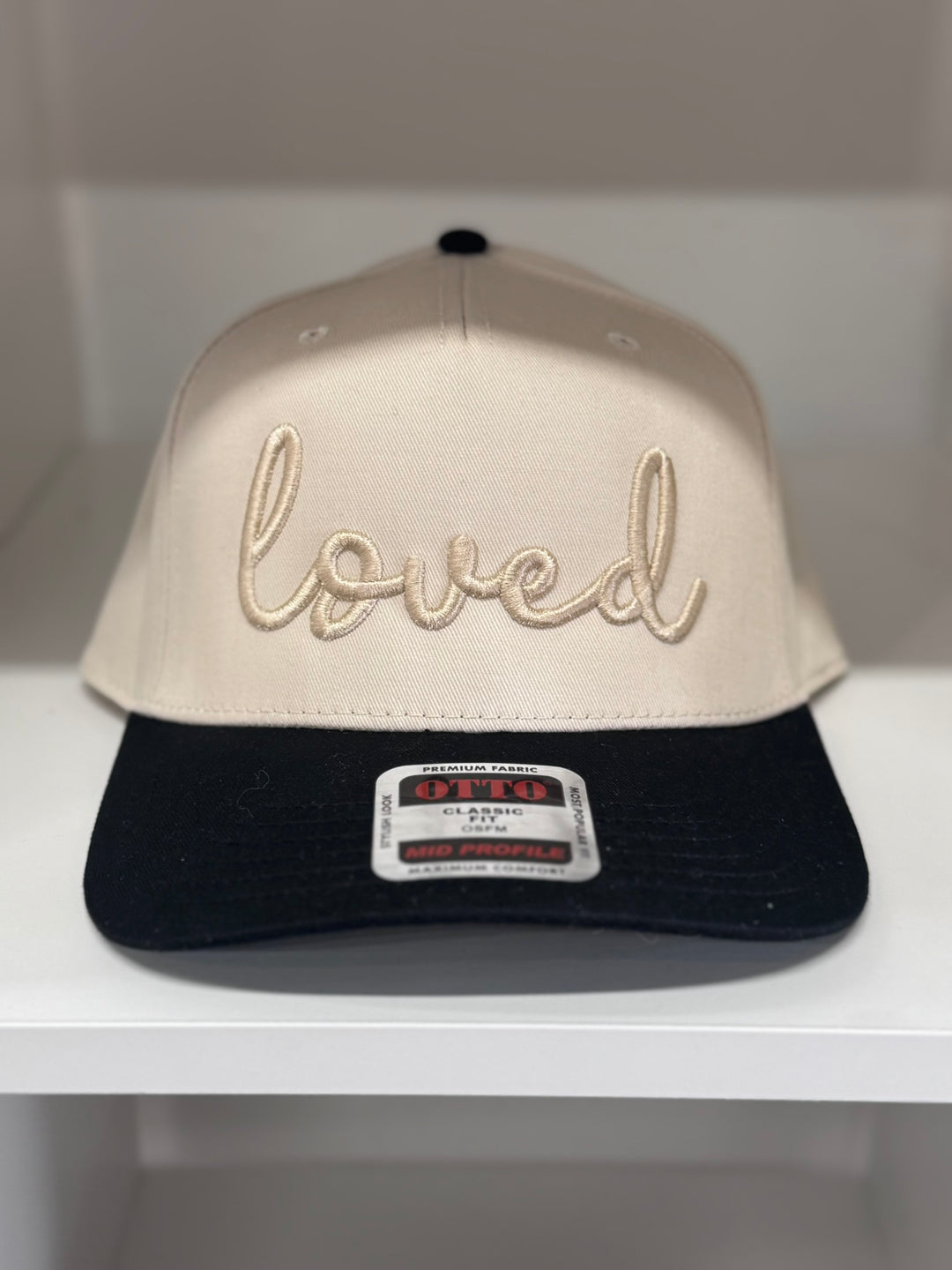 A tan baseball cap with "loved" embroidered in cursive on the front and a black bill.