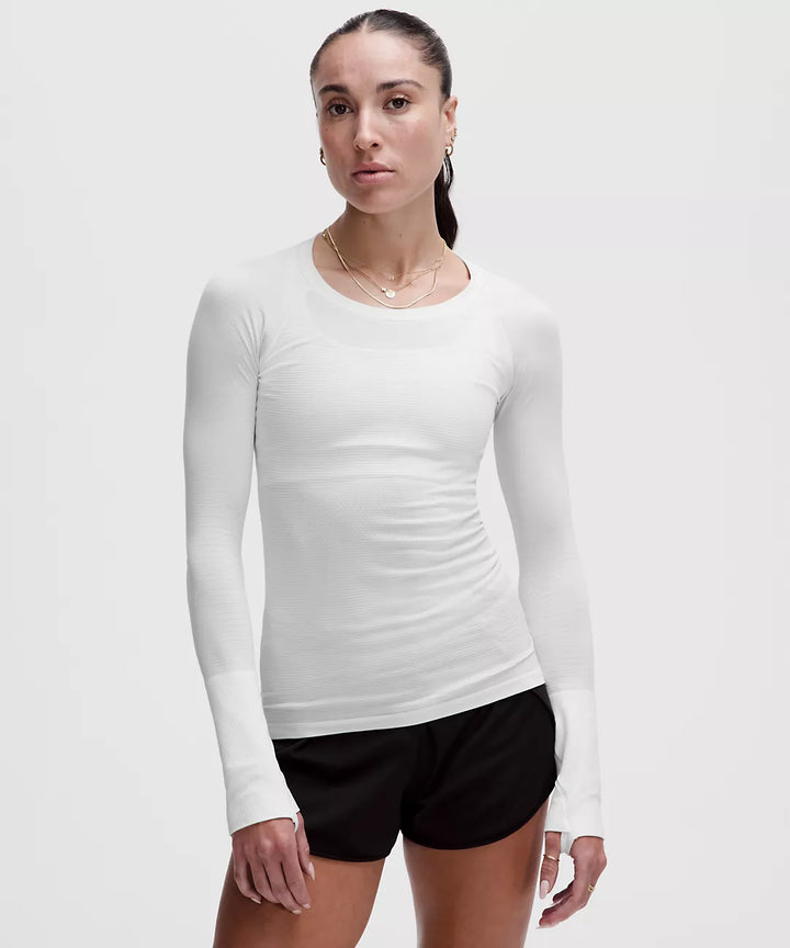 A woman wears a white long-sleeve athletic shirt with thumbholes and black shorts.
