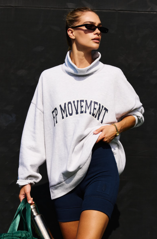 An athletic woman wears a grey oversized sweatshirt with "FP MOVEMENT" and navy biker shorts.