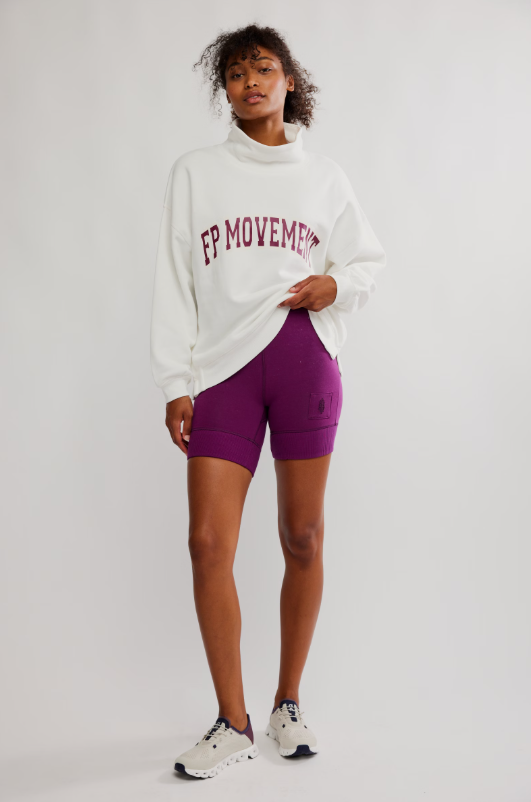 A woman models a white oversized sweatshirt with a funnel neck and purple biker shorts.