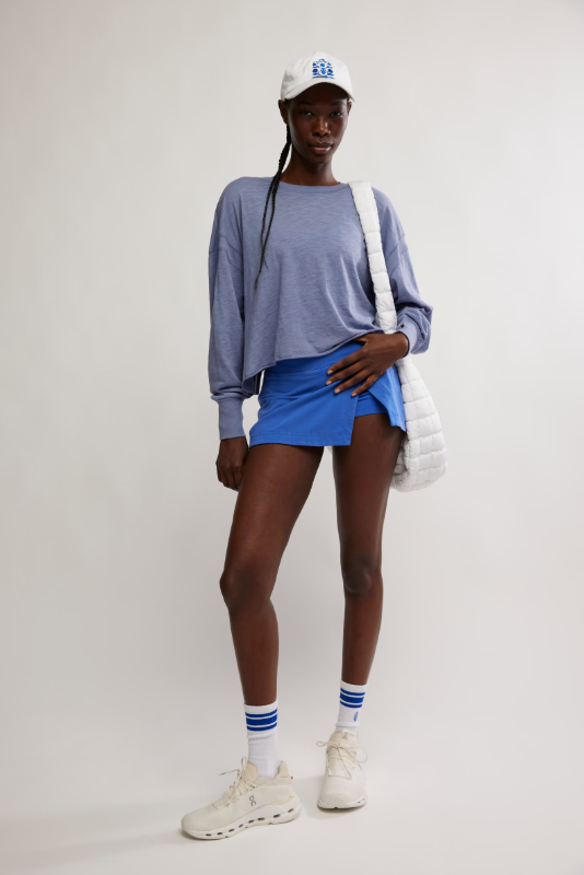 A woman wears a blue long-sleeve top, blue skort, white cap, and white sneakers.