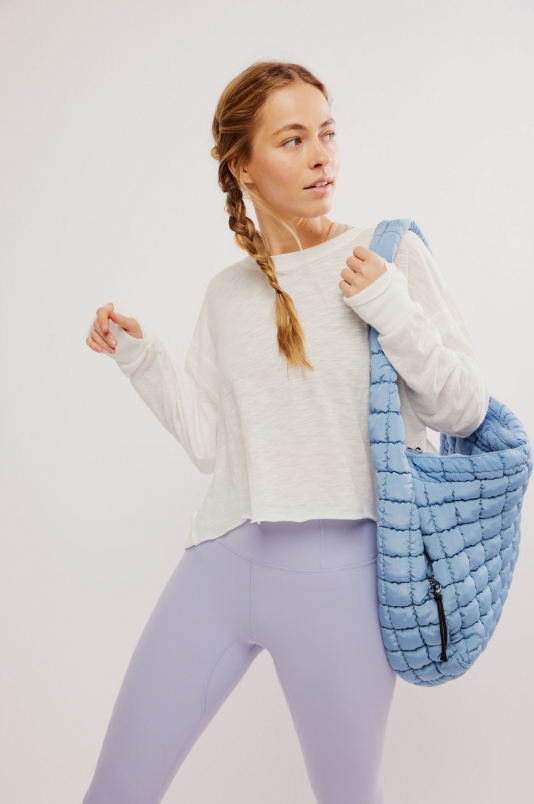 A woman in a white long-sleeve top and purple leggings holds a blue quilted bag.