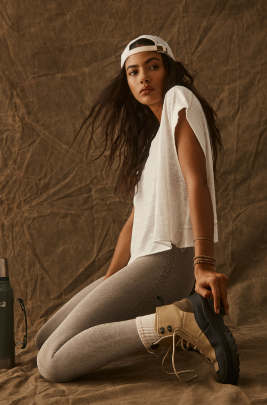 A woman in a white tee, grey leggings, and hiking boots kneels on a brown backdrop.
