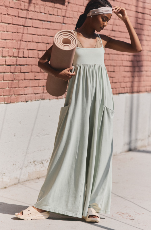 A woman wears a light green strappy onesie with wide legs and pockets, holding a yoga mat.