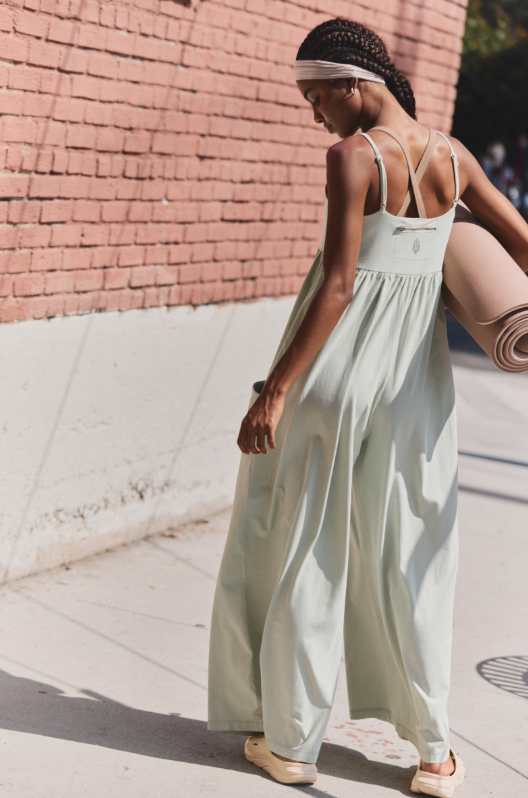 A woman in a light green strappy onesie with wide legs carries a yoga mat.