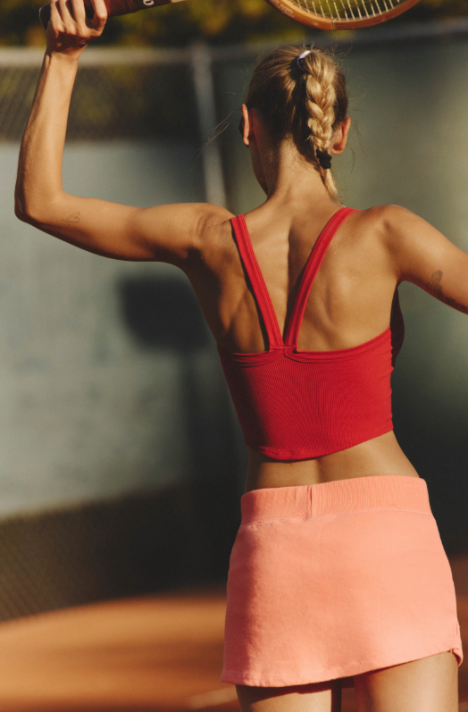 A woman from behind wears a cranberry cami and a peach skirt while playing tennis.