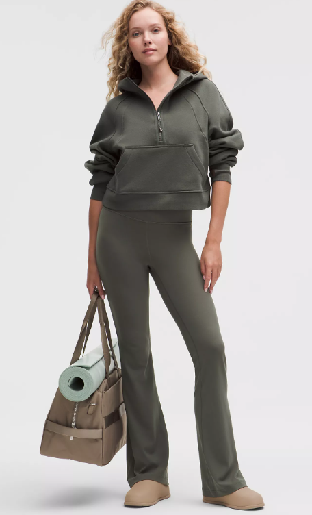 A woman wears a green half-zip hoodie and matching flared leggings, holding a gym bag with a yoga mat.