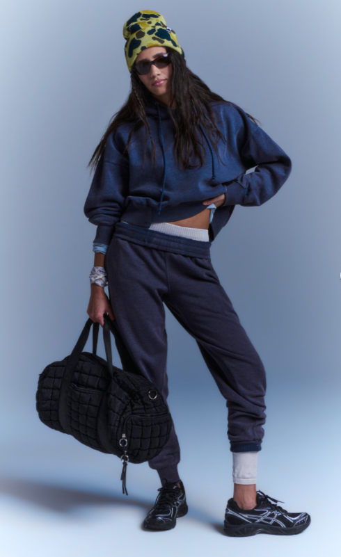 A woman wears a dark navy sand washed hoodie and sweatpants set with a patterned beanie and black sneakers.