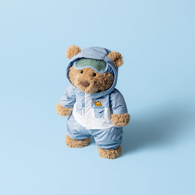 A brown teddy bear wears a light blue snowsuit with a hood and green goggles.