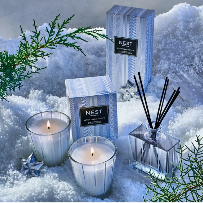 Two lit candles, a reed diffuser, and two boxes of Blue Cypress & Snow products sit in snow.