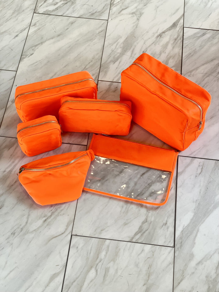 Five orange nylon pouches with gold zippers are arranged on a marble floor.