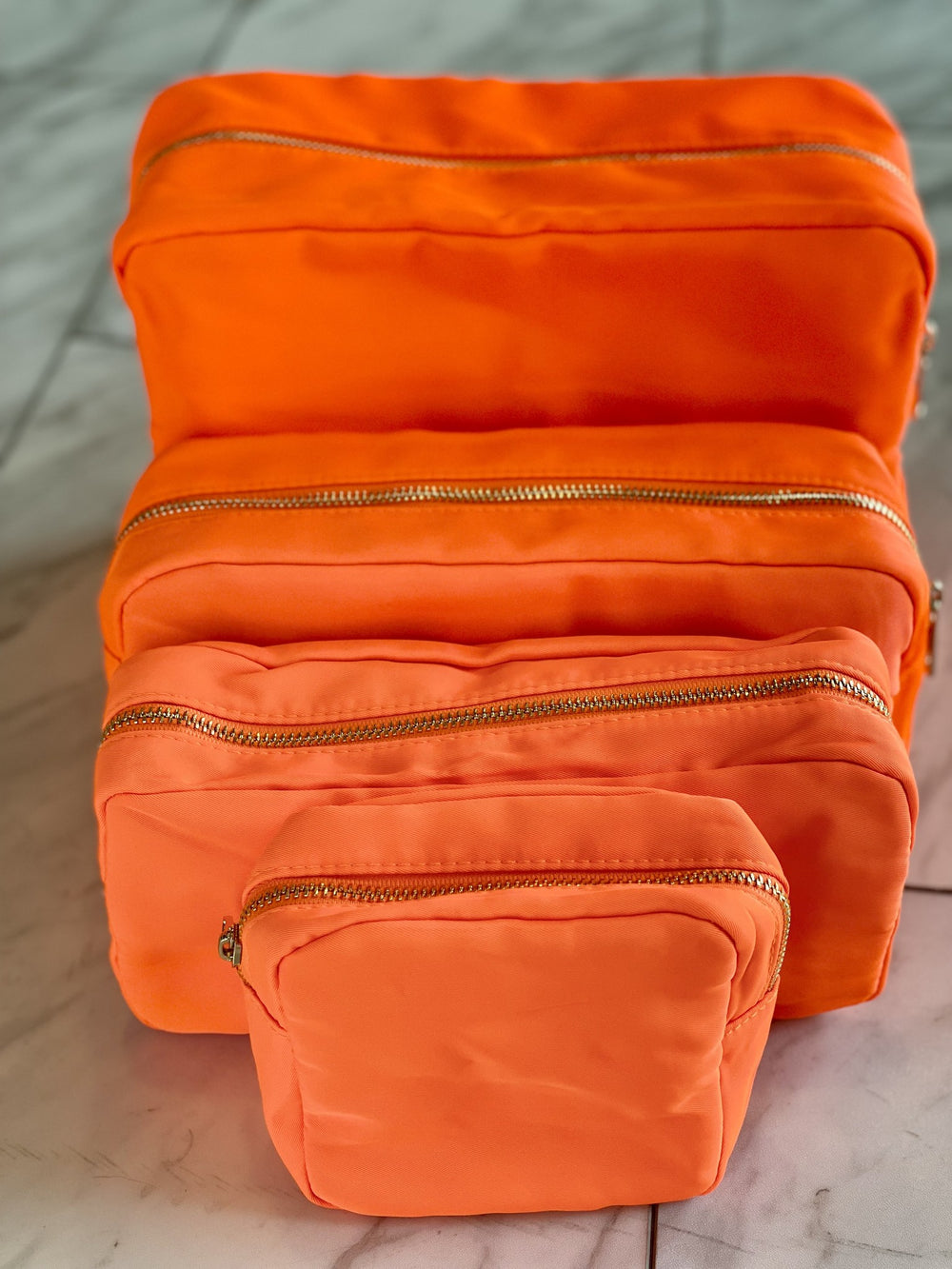 Four orange nylon pouches of varying sizes, each with a gold zipper, are stacked on a marble surface.