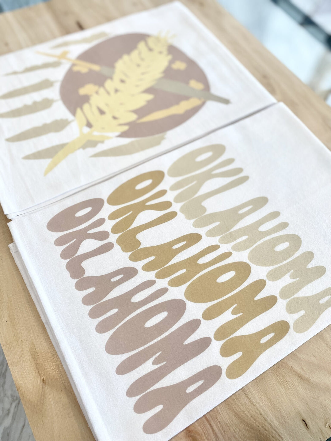 Two neutral-toned Oklahoma tea towels are stacked on a light wooden surface.