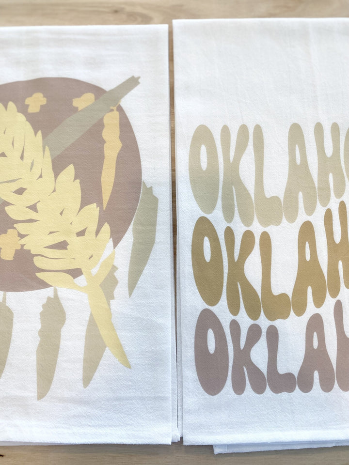 Two white tea towels are displayed, one with a neutral Oklahoma flag design and the other with "OKLAHOMA" in varying brown shades.