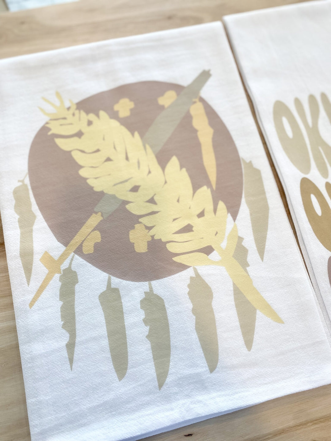 A white tea towel features a neutral-toned Oklahoma flag design with a feather and chili peppers.