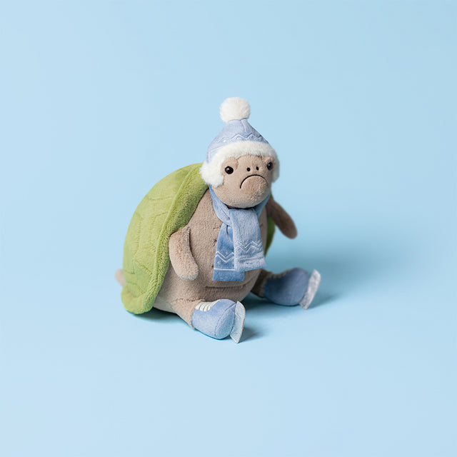 A plush turtle wearing a blue winter hat and scarf sits on a light blue background.